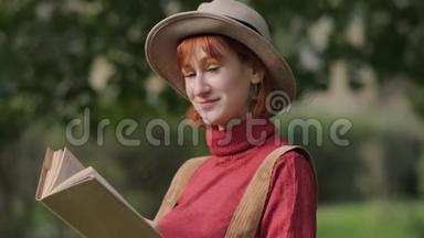 年轻迷人的红头发女人戴着帽子和毛衣在一个自然公园看书。 秋<strong>天凉</strong>爽的一<strong>天</strong>。